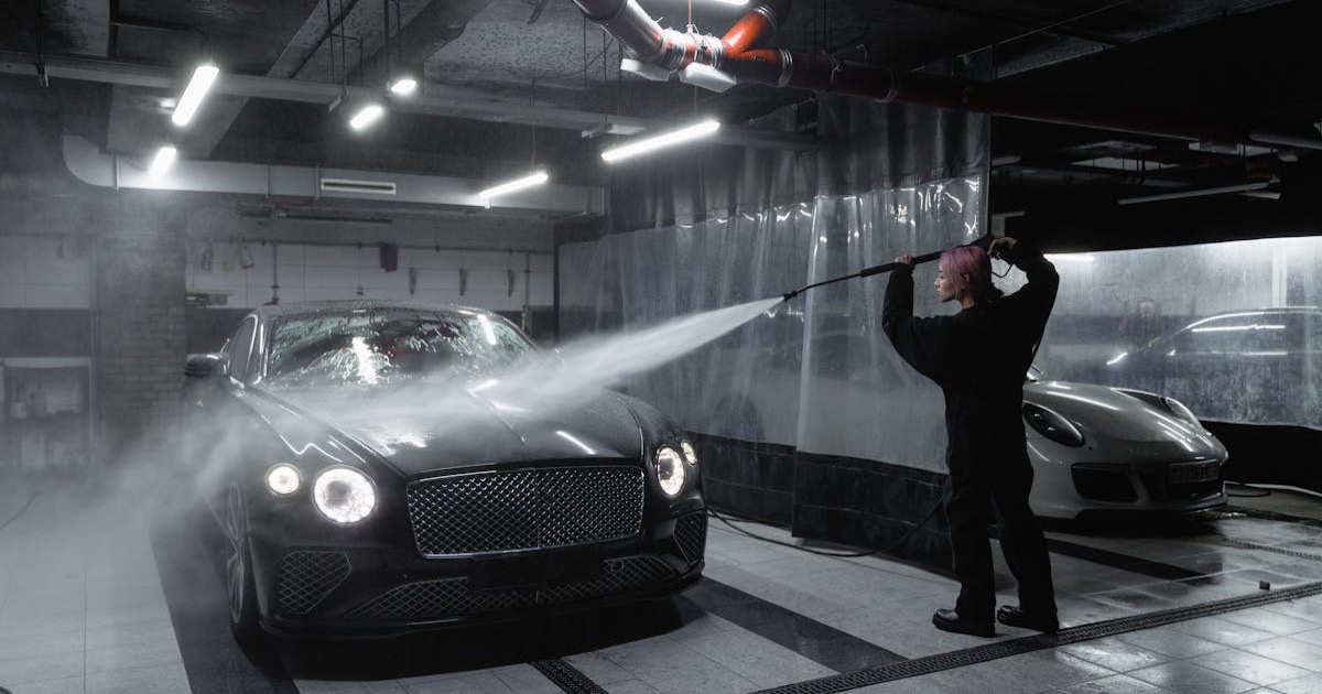 A person washing a black luxury car in a dimly lit garage using a pressure washer.