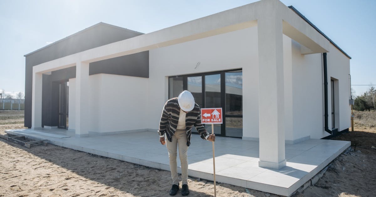 Real estate agent with hard hat standing by a modern house for sale.